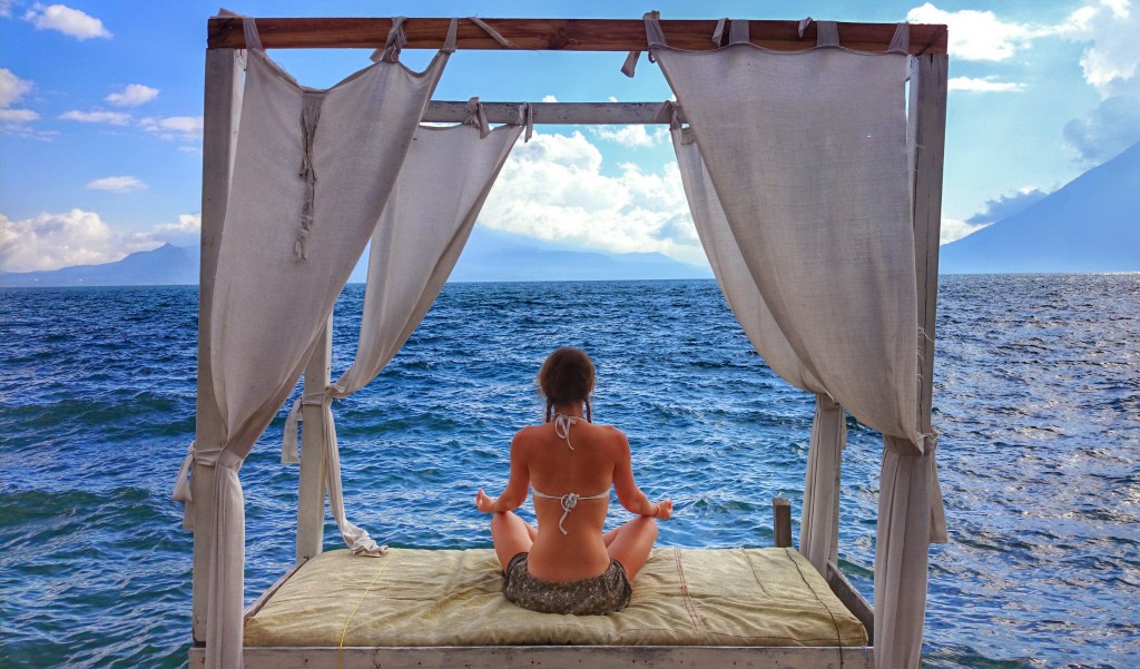 Image shows Jade sitting on a four poster bed cross legged and meditating, looking out towards the sea.